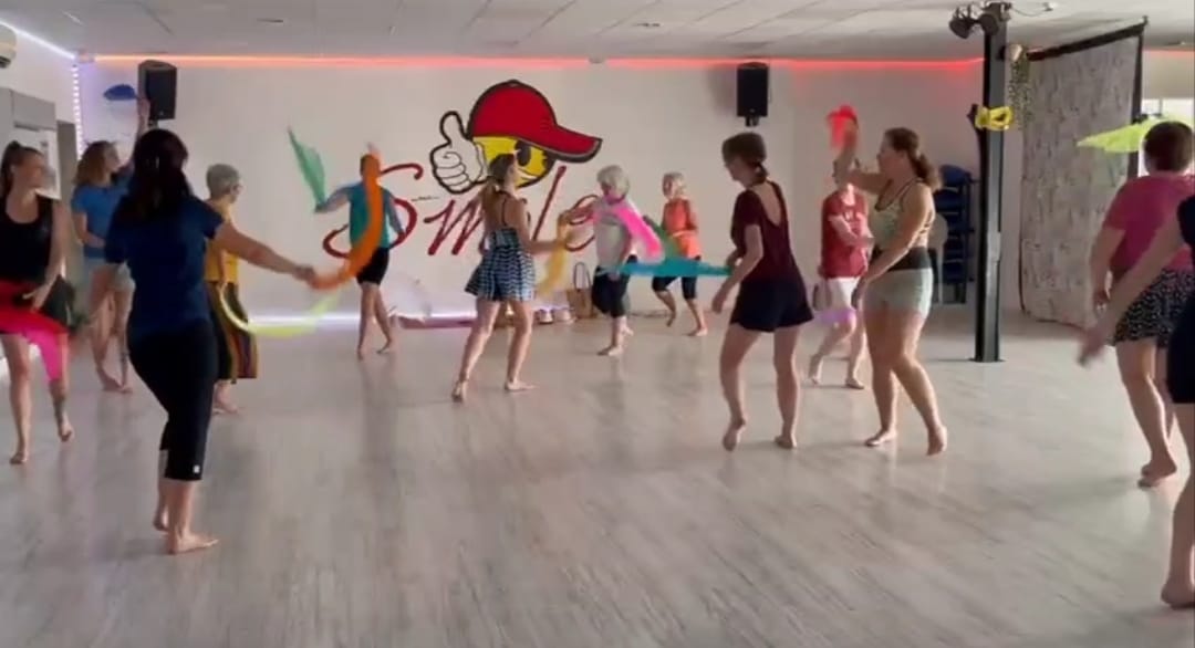 Cette photo montre un groupe de participants évoluant librement dans une salle de danse lumineuse. Chacun se déplace à son rythme dans l’espace, certains utilisant des rubans colorés qui prolongent les mouvements et renforcent la dimension ludique et créative de l’activité. Les participants sont pieds nus, favorisant une connexion directe avec le sol et les sensations corporelles. L’ambiance paraît détendue et conviviale, illustrant parfaitement l’esprit de la danse libre intuitive, où l’expression personnelle prime sur la technique ou la performance.