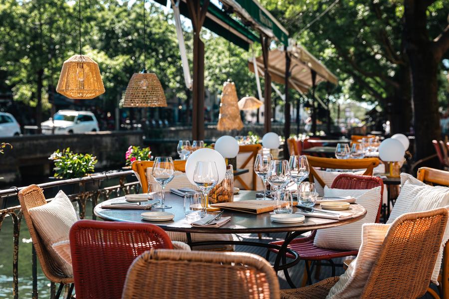 Cette vue met en valeur la terrasse de Monsieur TU, idéalement installée au bord de la Sorgue. Les tables soigneusement dressées, le mobilier élégant en fibres tressées et les suspensions naturelles créent une ambiance à la fois chic et décontractée. À l’ombre des arbres qui bordent la rivière, les convives profitent d’un cadre paisible et verdoyant. Une atmosphère propice à un déjeuner ensoleillé ou à un dîner romantique au fil de l’eau, dans l’un des plus beaux environnements de L’Isle-sur-la-Sorgue.