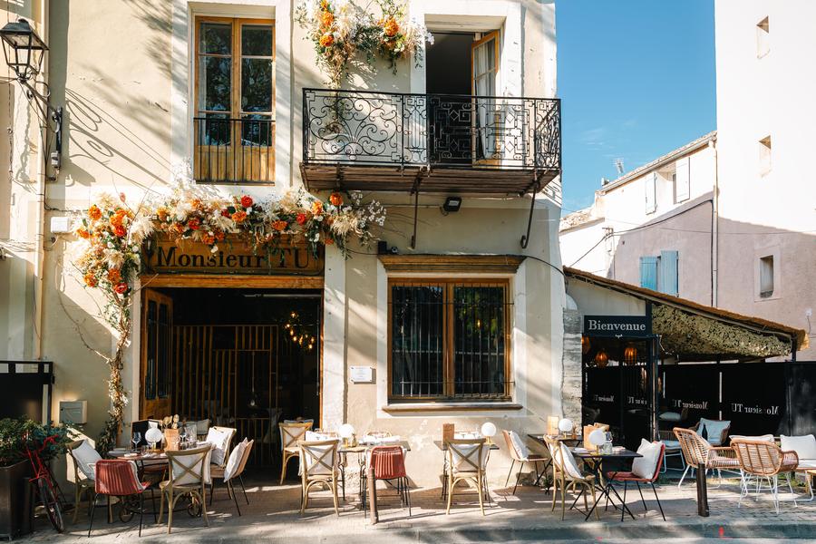 La façade de Monsieur TU dévoile une adresse pleine de charme au cœur de L’Isle-sur-la-Sorgue. Installé dans une élégante bâtisse de caractère aux tons clairs, le restaurant se distingue par ses compositions florales généreuses qui encadrent l’entrée et le balcon. Quelques tables dressées en terrasse invitent à profiter de l’ambiance animée des quais, tandis que les grandes ouvertures laissent entrevoir une atmosphère chaleureuse à l’intérieur. L’ensemble évoque une adresse raffinée et conviviale, typique de l’art de vivre provençal.