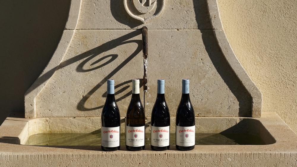Sélection de bouteilles du Clos du Calvaire présentées devant une fontaine en pierre provençale. Cette mise en scène illustre les différentes cuvées élaborées par le domaine au cœur de l'appellation Châteauneuf-du-Pape.