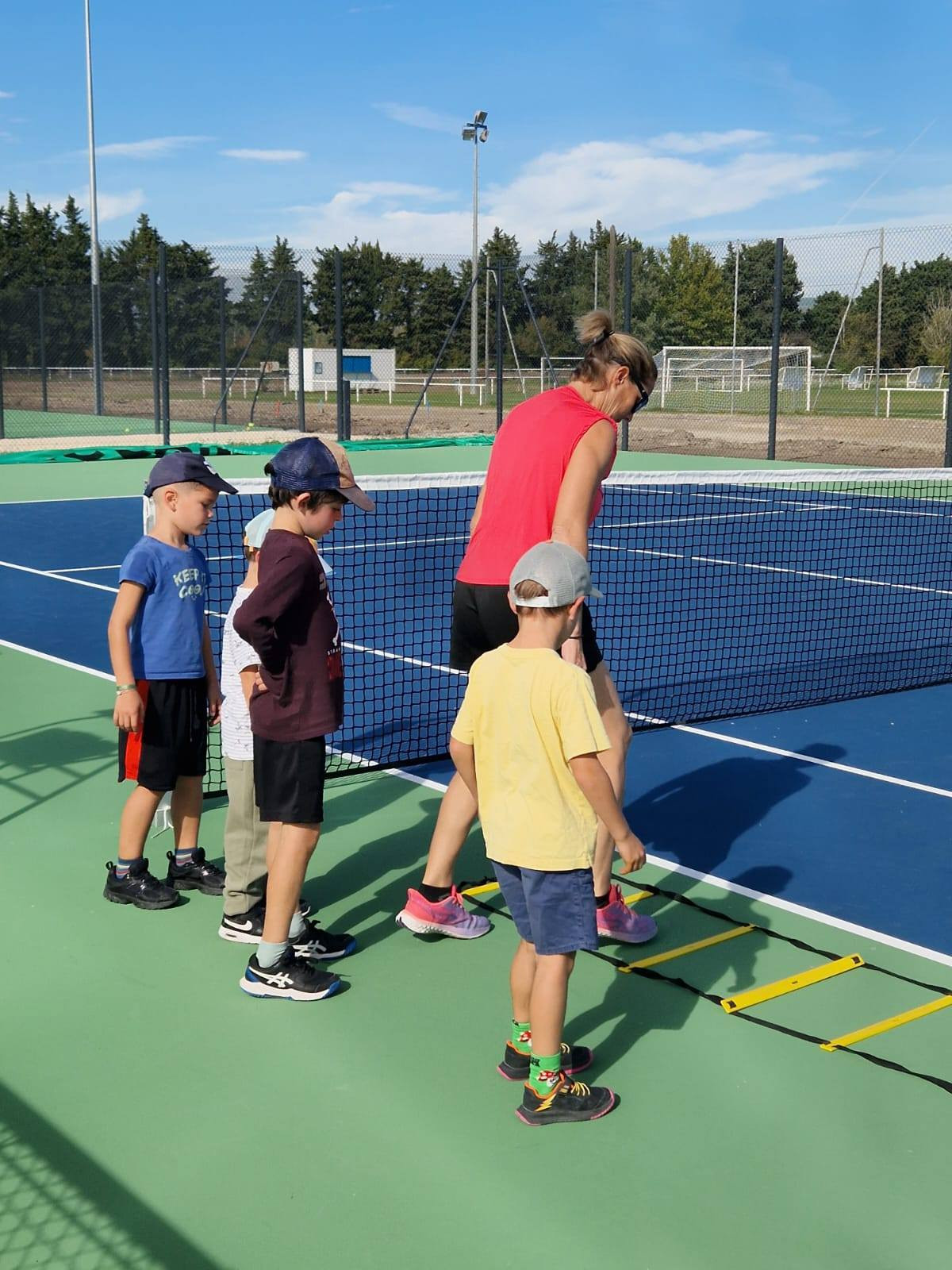 **Description :** Une éducatrice encadre un groupe de jeunes enfants lors d'une séance d'initiation au tennis sur un court extérieur. À l'aide d'une échelle de rythme posée au sol, les participants réalisent un exercice de coordination et de motricité dans une ambiance ludique et conviviale. Les terrains modernes et le complexe sportif se distinguent en arrière-plan sous un ciel dégagé.
