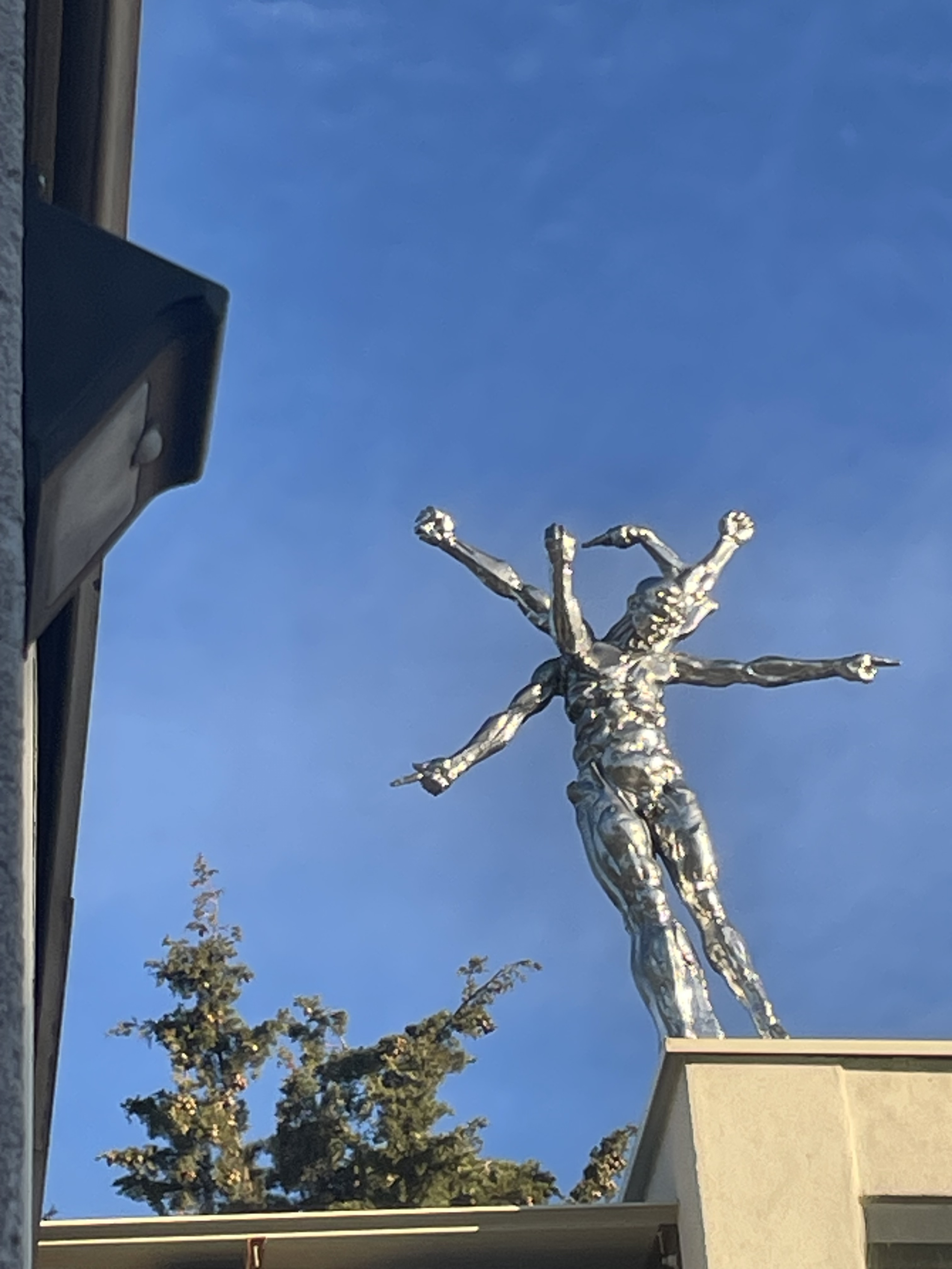 Cette photographie met en valeur une sculpture monumentale représentant une figure humaine aux multiples bras, réalisée dans une finition métallique brillante qui capte la lumière. Installée en hauteur sur un bâtiment, l'œuvre se détache sur un ciel bleu profond et semble défier les lois de l'équilibre. Les différentes positions des bras suggèrent le mouvement, l'élan et la multiplicité des actions, conférant à la sculpture une impression de dynamisme et de liberté.