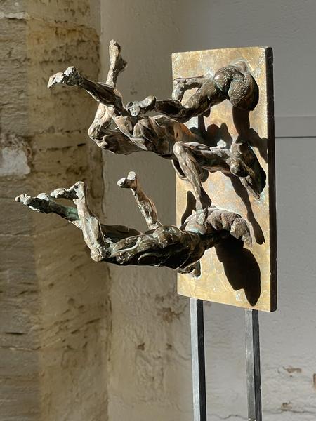 Cette seconde image présente une sculpture en bronze composée de deux figures humaines semblant émerger d'une plaque verticale. Les corps, aux formes expressives et puissantes, semblent suspendus dans un mouvement de bascule ou d'envol, créant un dialogue entre la matière, le vide et l'espace. Les jeux de lumière révèlent la texture du bronze et soulignent la force du modelé, illustrant le travail de Vincent Liévore autour du mouvement, de l'équilibre et de la relation entre la sculpture et son support.