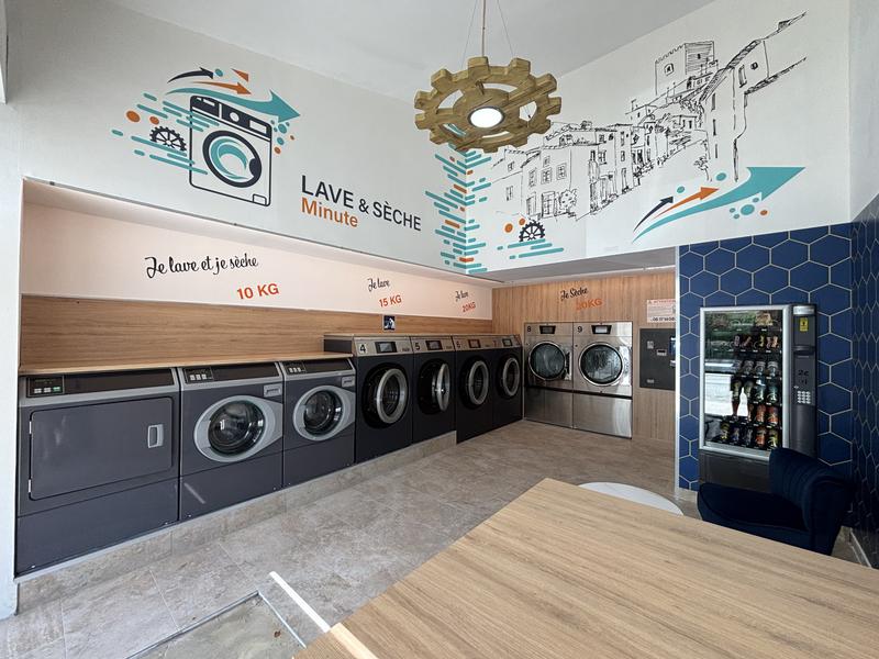 Vue d'ensemble de la laverie automatique Lave & Sèche Minute à L'Isle-sur-la-Sorgue. L'espace moderne accueille plusieurs machines à laver et sèche-linge de différentes capacités, ainsi qu'un distributeur automatique de produits et de boissons. La décoration met en avant l'identité locale avec une fresque murale inspirée du patrimoine de la ville.