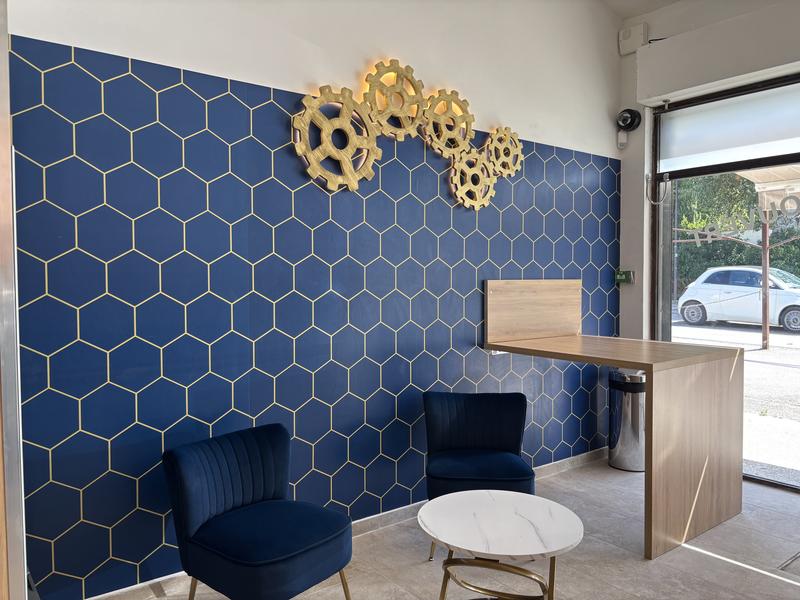 L'espace d'accueil de Lave & Sèche Minute offre un cadre confortable avec des fauteuils, une table d'attente et un comptoir. La décoration contemporaine, composée d'un mur bleu à motifs géométriques et d'éléments évoquant les roues à aubes de L'Isle-sur-la-Sorgue, crée une ambiance agréable pendant le temps de lavage ou de séchage.