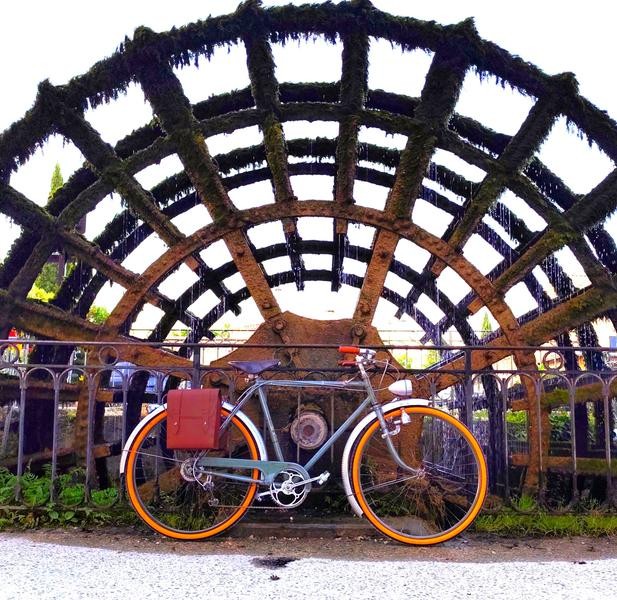 Un vélo de ville ancien noir est photographié devant une roue à aubes, symbole du patrimoine de L'Isle-sur-la-Sorgue. L'image évoque le lien entre histoire, nature et découverte de la destination.