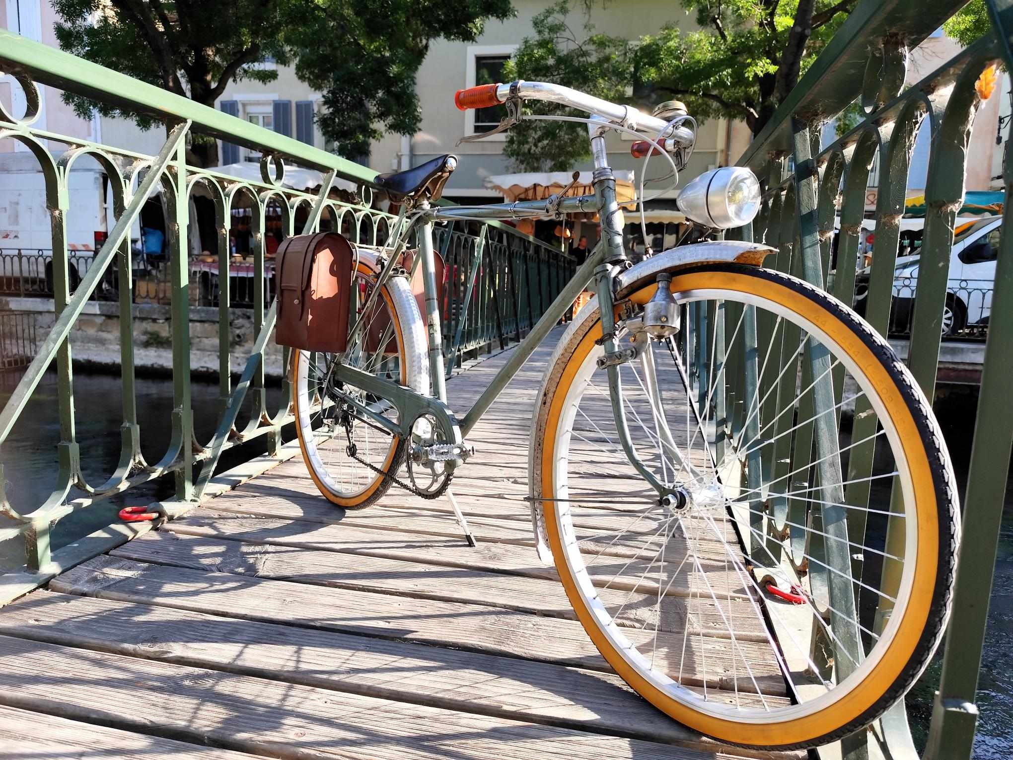 Un vélo vintage est photographié sur une passerelle métallique traversant la Sorgue. Le décor met en avant les canaux emblématiques de la ville et invite à une découverte à vélo.