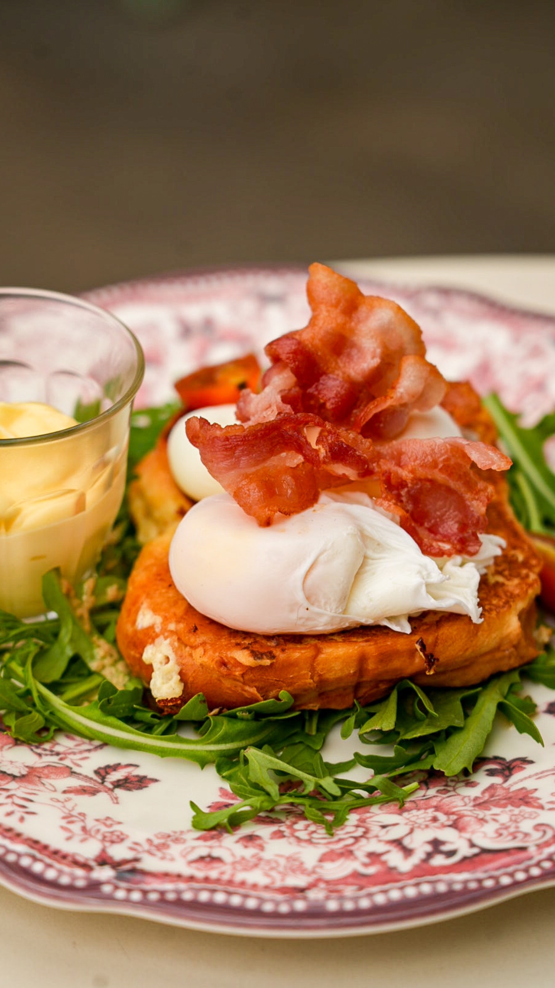 Une généreuse tranche de brioche dorée accueille deux œufs pochés au cœur fondant, délicatement surmontés de tranches de bacon croustillant. L'ensemble repose sur un lit de roquette fraîche et s'accompagne d'une sauce onctueuse servie à part, idéale pour napper le plat selon les envies. Présentée dans une assiette aux motifs fleuris, cette assiette mêle gourmandise et élégance, illustrant parfaitement l'esprit d'un brunch généreux et fait maison.