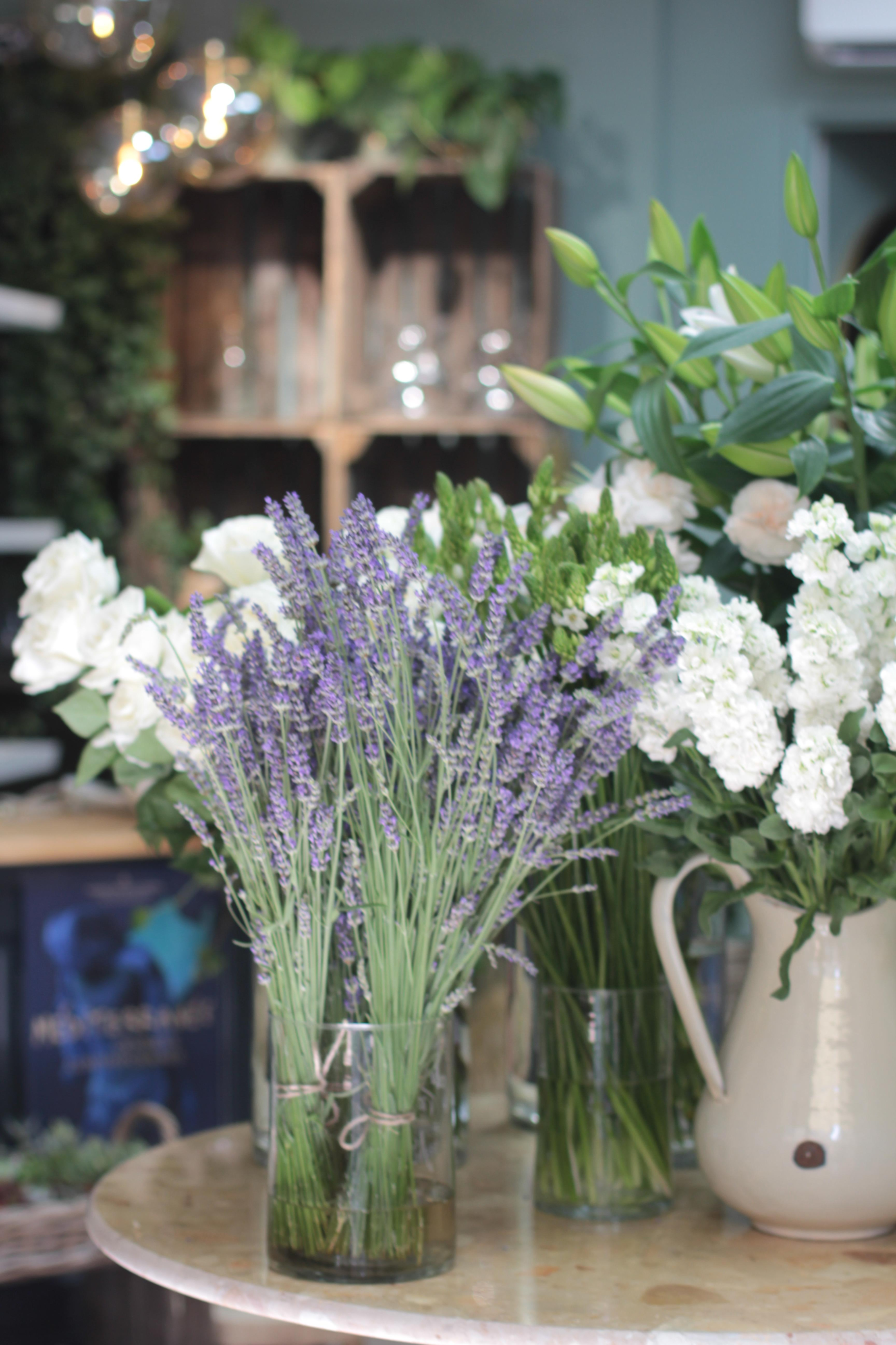 Présentation de fleurs fraîches en boutique, avec au premier plan plusieurs bouquets de lavande. Ils sont entourés de compositions blanches, notamment de lys et d’hortensias, dans une ambiance végétale et authentique.
