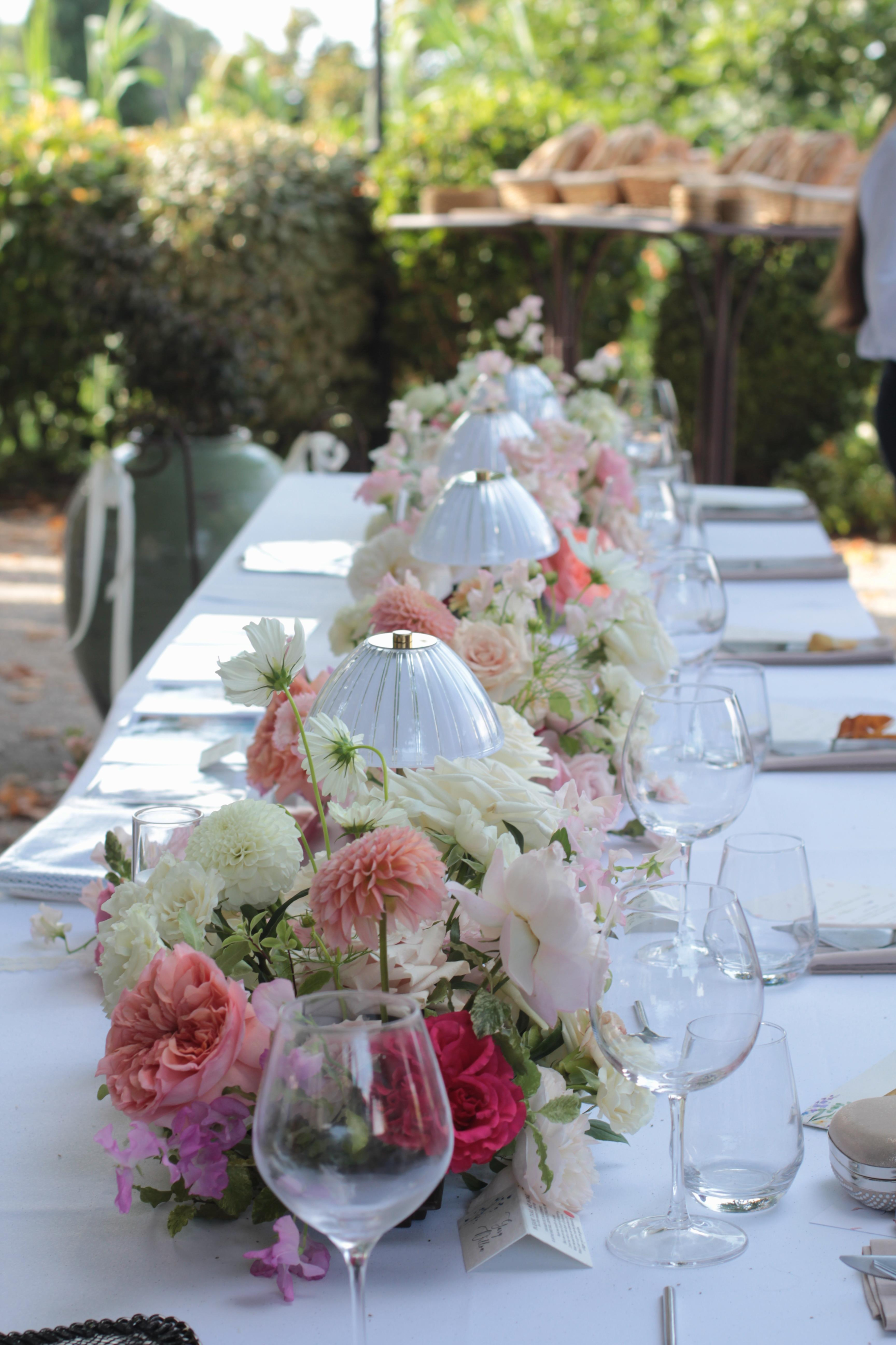 Longue table de réception dressée en extérieur, décorée d’un généreux chemin de fleurs dans des tons blanc, rose poudré et fuchsia. Des verres, de petites lampes de table et une vaisselle élégante composent une atmosphère raffinée, idéale pour un mariage ou une réception.