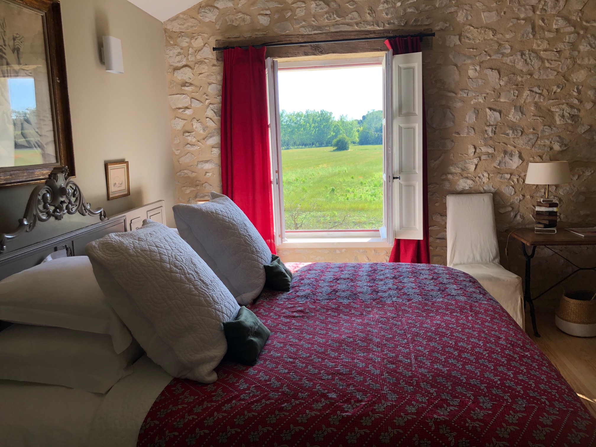 Chambre de charme du Mas de la Sorgue avec murs en pierre apparente, linge de lit aux tons rouges et grande fenêtre ouverte sur la campagne verdoyante.