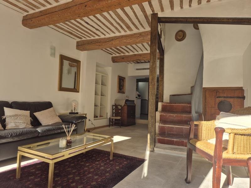 Salon chaleureux avec poutres apparentes, plafond traditionnel, mobilier ancien et contemporain, canapé et fauteuils. L’escalier en tomettes et les éléments en bois renforcent le charme authentique de cette maison de village.