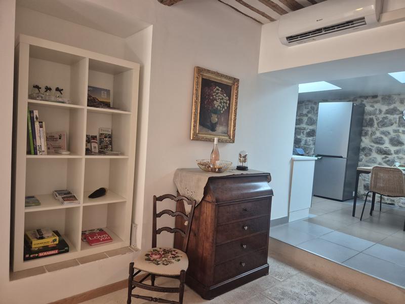 Vue sur un espace de vie mêlant décoration traditionnelle et équipements contemporains, avec un secrétaire ancien, une bibliothèque intégrée et une cuisine ouverte aux murs en pierre. La pièce est équipée de la climatisation et conserve plusieurs éléments de caractère.