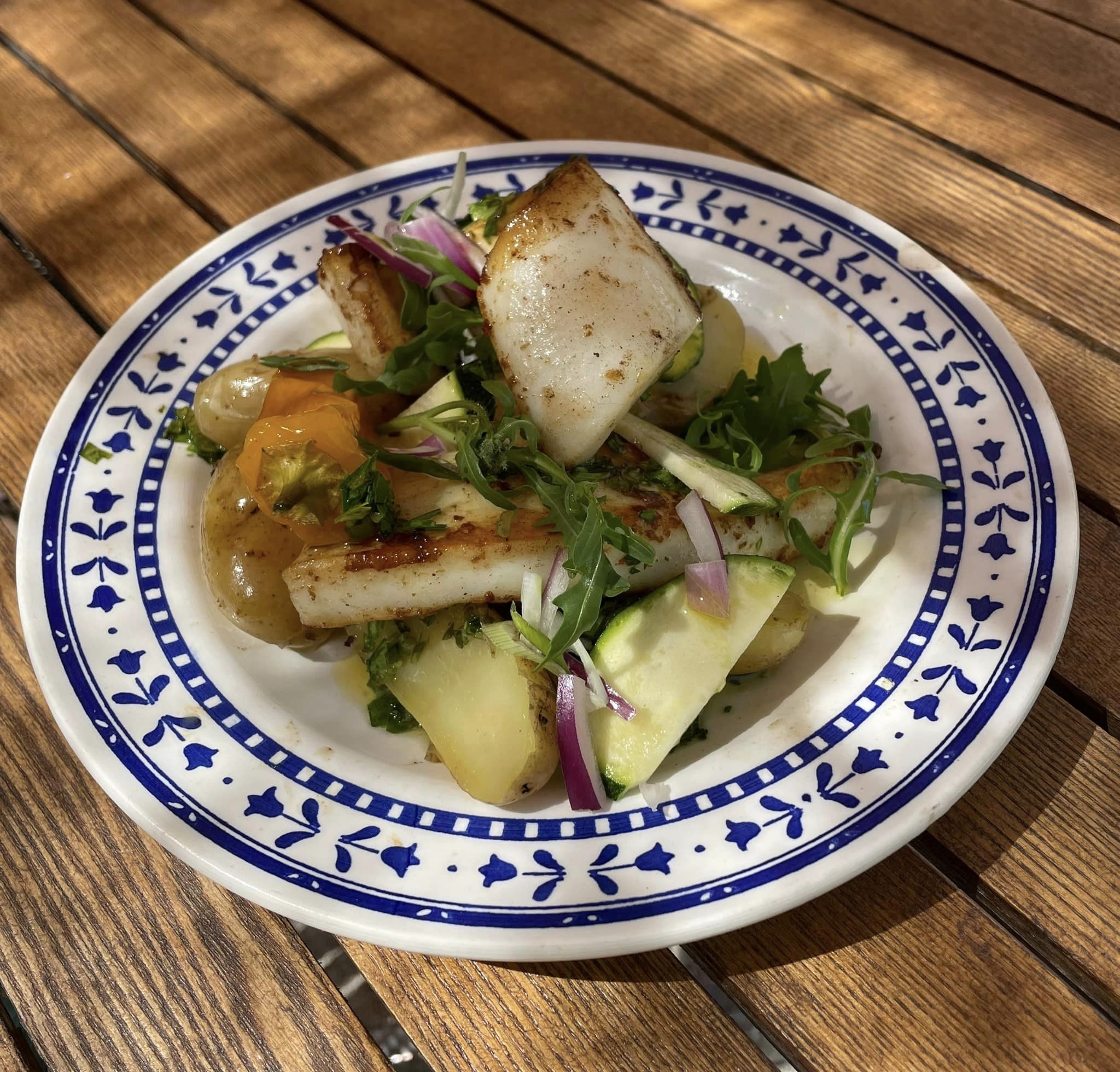 Assiette de salade fraîche avec des morceaux de seiche grillée, des pommes de terre, des courgettes, des oignons rouges, de la roquette et des herbes, posée sur une table en bois.