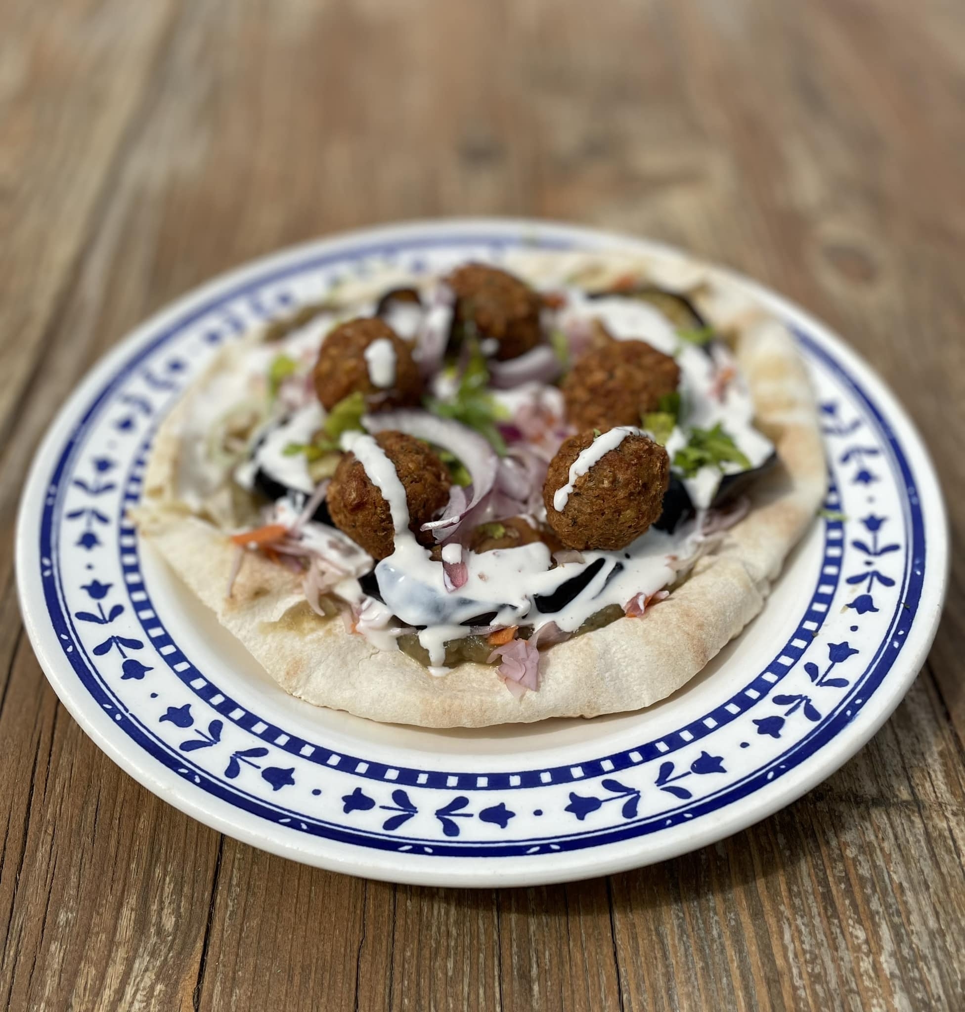 Assiette de falafels ouverts sur un pain pita, garnie d'oignons rouges, d'aubergines, de chou râpé et arrosée de sauce blanche, posée sur une table en bois.