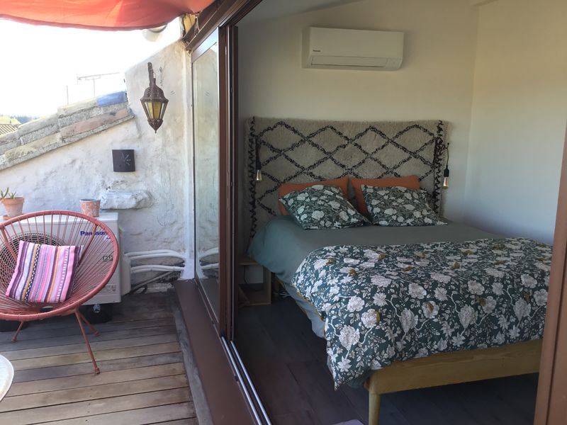 Chambre lumineuse avec un lit double orné d'une tête de lit en tissu à motif géométrique et d'une literie florale, s'ouvrant sur une terrasse en bois équipée d'un fauteuil et de plantes, sous un parasol rouge.