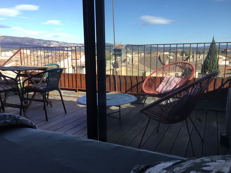 Terrasse aménagée avec des meubles d'extérieur variés, incluant des chaises en rotin et des fauteuils modernes, offrant une vue panoramique sur un village aux toits de tuiles et des collines en arrière-plan sous un ciel bleu.