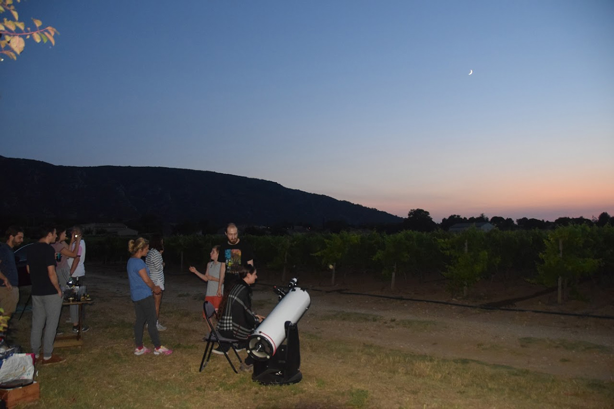 Un groupe de personnes observe le ciel nocturne au coucher du soleil dans un champ avec un télescope blanc au premier plan. Une colline sombre et une vigne sont visibles en arrière-plan sous un ciel passant du bleu au rose et un croissant de lune.