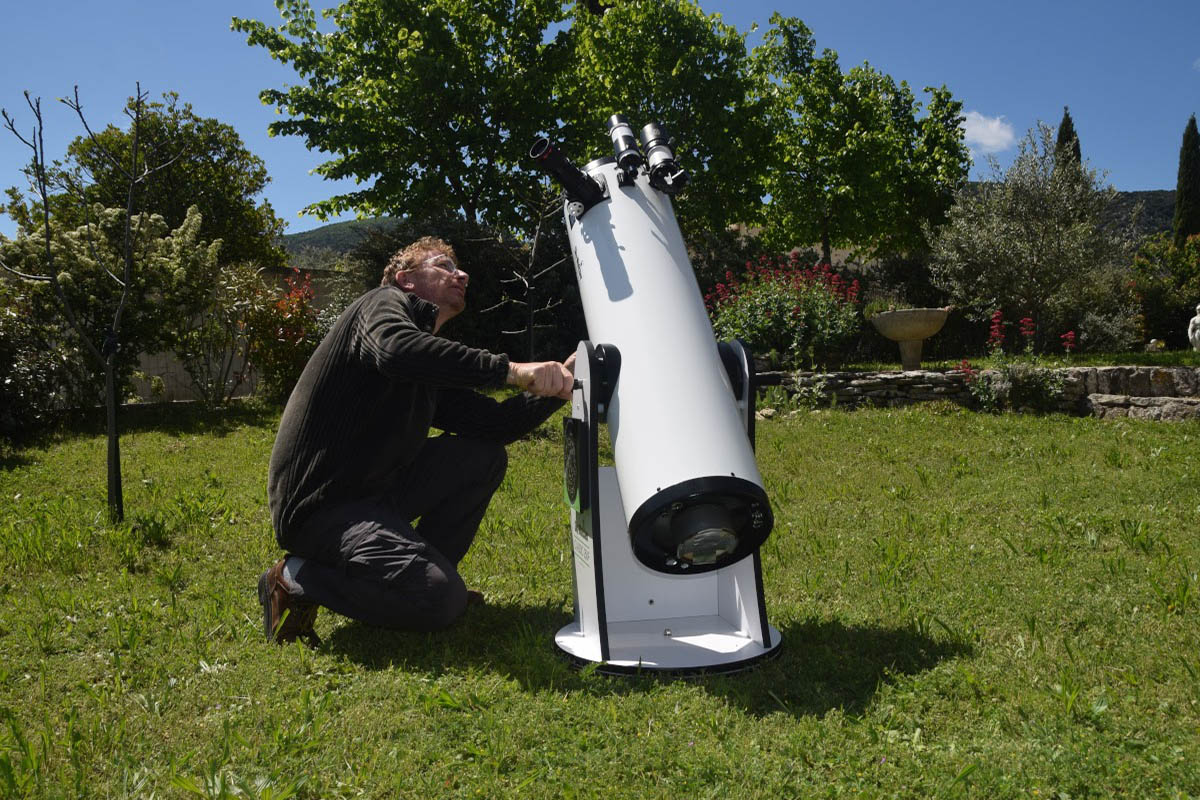 Un homme accroupi ajuste un grand télescope blanc de type Dobson, placé sur l'herbe d'un jardin. Des arbres verts et des fleurs sont visibles en arrière-plan sous un ciel bleu.