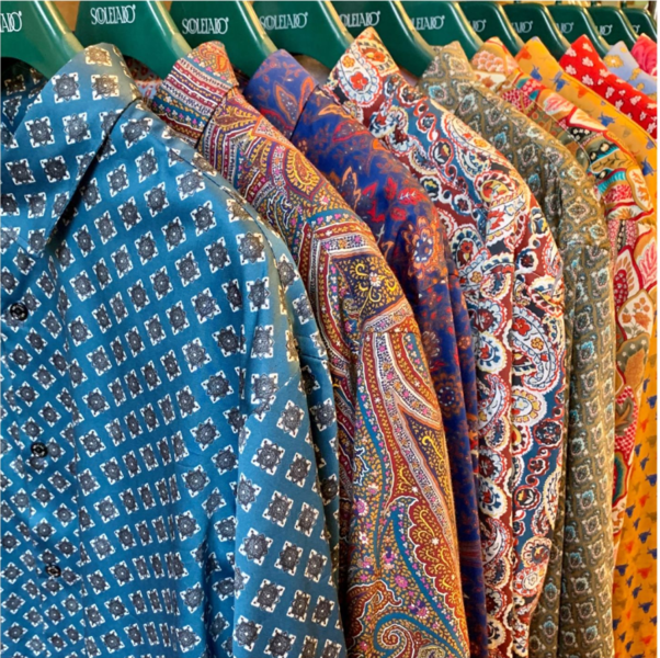 Une série de chemises à motifs colorés sont suspendues sur des cintres, alignées dans une boutique. Les motifs varient, allant de motifs géométriques bleus à des motifs cachemire plus élaborés dans des tons rouges, orange et bleus.