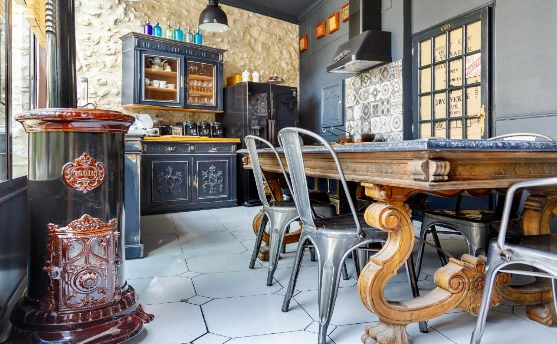 Une salle à manger au style rustique et industriel, avec une cheminée à bois marron foncé au premier plan à gauche. Un grand meuble en bois sombre avec des vitrines est visible au fond. Sur la droite, une longue table en bois ornée est entourée de chaises en métal. Les murs sont un mélange de pierre apparente et de peinture grise, et le sol est carrelé de blanc. Au fond, une hotte de cuisine est installée au-dessus d'un carrelage mural décoratif.