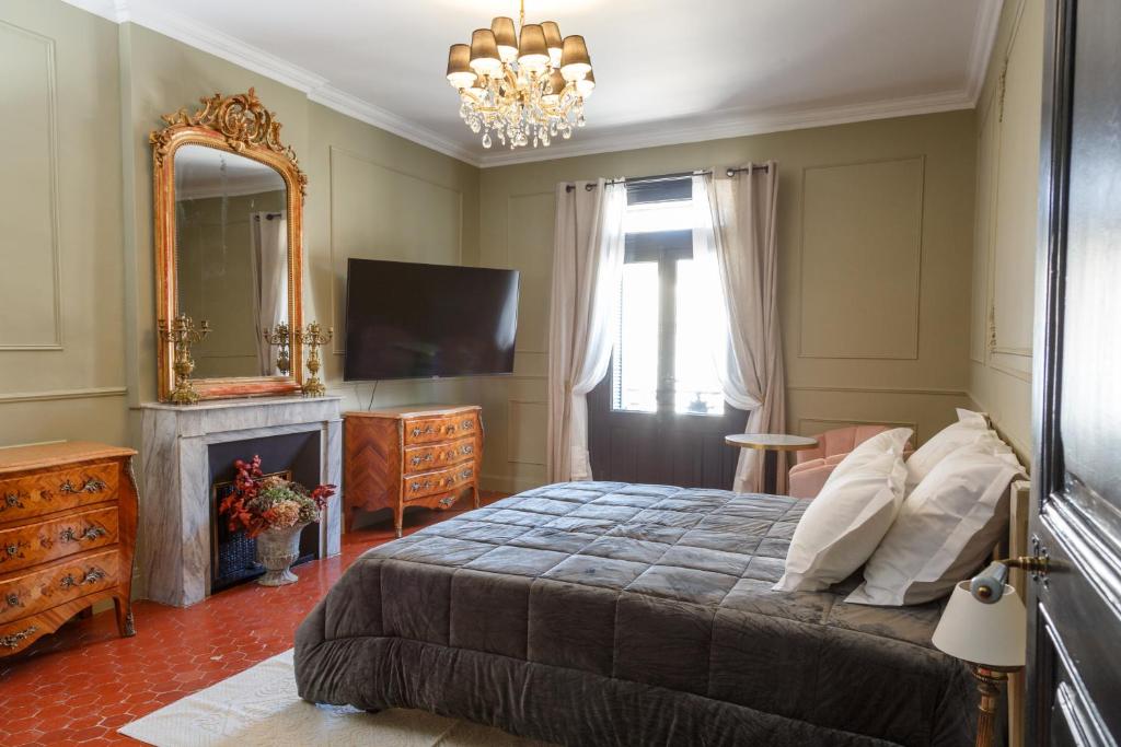 Une chambre luxueuse avec un grand lit king-size drapé d'une couette grise. Au-dessus du lit, un lustre doré et en cristal illumine la pièce. Un grand miroir doré orné est posé sur une cheminée en marbre. En face du lit, une télévision à écran plat est fixée au mur. Deux commodes en bois ornées sont visibles de chaque côté de la pièce. À droite, une fenêtre encadrée de rideaux clairs apporte de la lumière naturelle. Le sol est recouvert de carrelage en terre cuite.