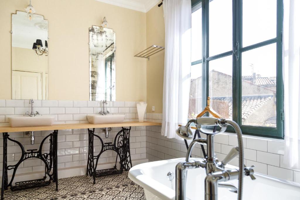 Une salle de bain lumineuse avec des murs crème, des carrelages blancs et un sol à motifs. Elle comprend une grande baignoire blanche avec un robinet en chrome au premier plan, et un long comptoir en bois avec deux lavabos rectangulaires, chacun reposant sur une base de machine à coudre ancienne en fer forgé. Deux grands miroirs anciens sont accrochés au-dessus des lavabos et une grande fenêtre à carreaux sombres est visible à droite, avec des rideaux blancs vaporeux.
