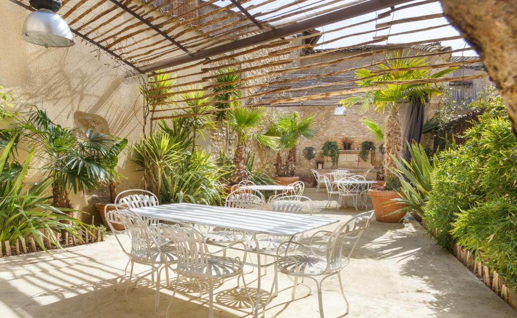 Une cour extérieure aménagée en salle à manger, avec une longue table blanche et des chaises assorties au premier plan. De nombreuses plantes vertes et des palmiers entourent la zone. Une structure de pergola en bois et en métal au-dessus offre de l'ombre, et une suspension métallique pend du côté gauche. D'autres petites tables rondes sont visibles au fond.