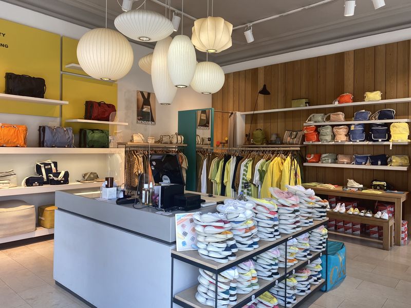 L'intérieur d'un magasin lumineux et bien rangé, avec des murs blancs et du bois clair. Des étagères blanches présentent une variété de sacs à main et de sacs à dos colorés, tandis que des portants sont remplis de vêtements. Un comptoir de caisse gris se trouve au centre, avec des piles de chaussures et des accessoires organisés sur des présentoirs.