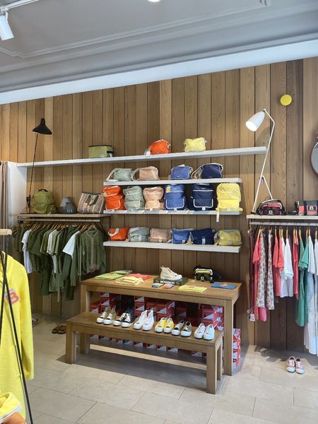 L'intérieur d'une boutique bien éclairée, avec un mur en bois clair et des étagères blanches remplies de sacs à main et de sacs à dos colorés. Des vêtements sont suspendus sur des portants en bois au premier plan, et une table en bois présente des chaussures et d'autres accessoires. Une lampe de sol blanche et une applique murale noire complètent le décor.