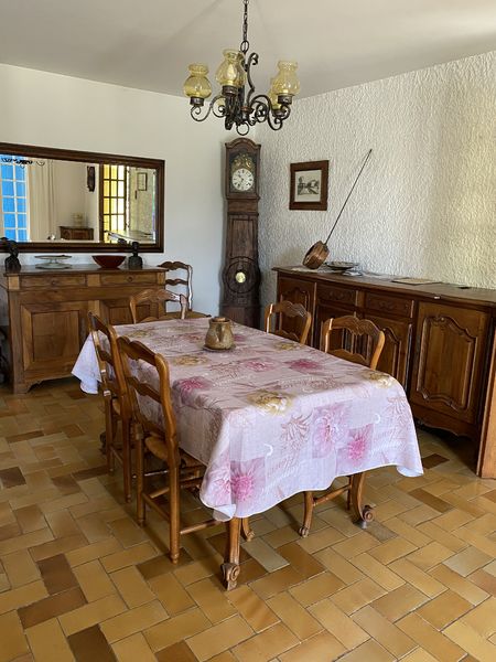 Une salle à manger lumineuse dans un style rustique, avec un sol carrelé et des murs blancs texturés. Au centre, une table en bois recouverte d'une nappe à motifs roses est entourée de chaises en bois. Deux grands buffets en bois sombre sont placés le long des murs, l'un avec un miroir horizontal au-dessus et l'autre avec une horloge grand-père à côté. Un lustre de style ancien pend du plafond.