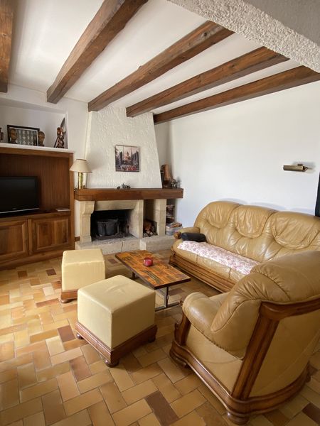 Une pièce à vivre chaleureuse de style rustique, dotée d'un sol en carreaux de terre cuite et de poutres apparentes au plafond. Au centre se trouvent un canapé et un fauteuil en cuir couleur camel, un meuble télé en bois, et une cheminée en pierre avec un manteau en bois. Deux poufs de couleur crème complètent le mobilier autour d'une table basse en bois.