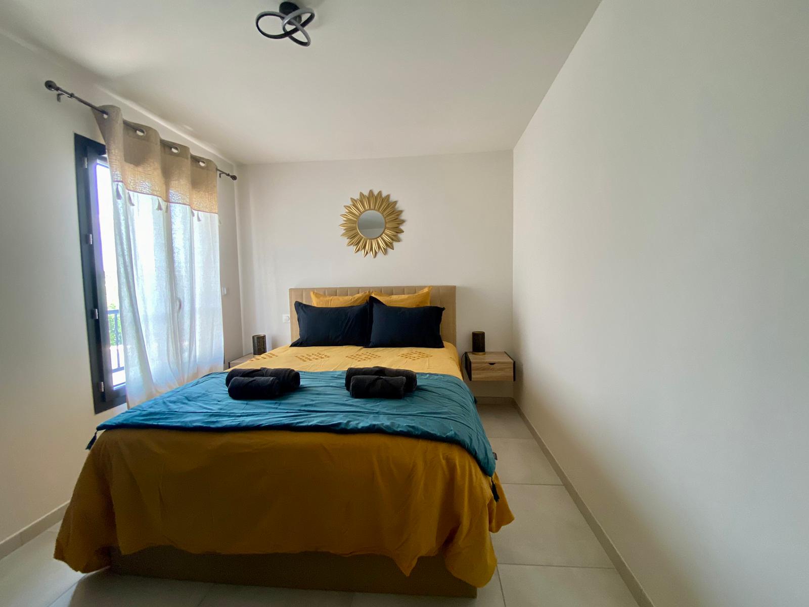     Requête réussie  Une chambre à coucher lumineuse avec un lit double orné d'une couette et de coussins blancs, un couvre-lit rayé blanc et gris, et un plaid à pompons. Une table de chevet en bois foncé avec une lampe moderne est visible à gauche.