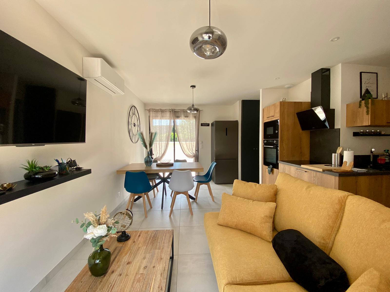 Un salon moderne et lumineux avec une grande télévision à écran plat noire fixée au mur. Un canapé jaune clair avec des coussins jaunes et un oreiller noir est visible au premier plan. Une table basse en bois est posée sur un carrelage blanc. En arrière-plan, une salle à manger avec une table en bois et des chaises colorées. Une cuisine ouverte avec des armoires en bois est également visible sur la droite.