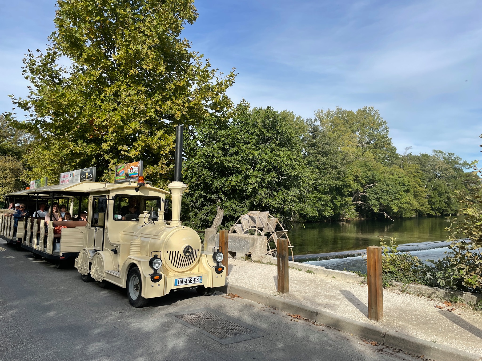 Un petit train touristique de couleur crème, avec un moteur en forme de locomotive et des wagons ouverts remplis de passagers, roule le long d'une route au bord d'une rivière. La berge est bordée d'arbres feuillus et d'une roue à aubes visible sur la droite. Le ciel est partiellement nuageux.