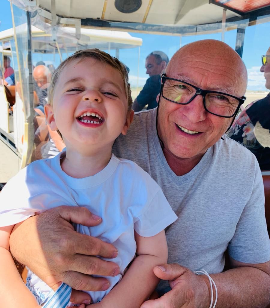 Un homme chauve avec des lunettes souriant et tenant un jeune enfant souriant et riant, tous deux semblant profiter de leur temps ensemble. Ils sont assis dans un véhicule ouvert, avec d'autres personnes floues en arrière-plan.