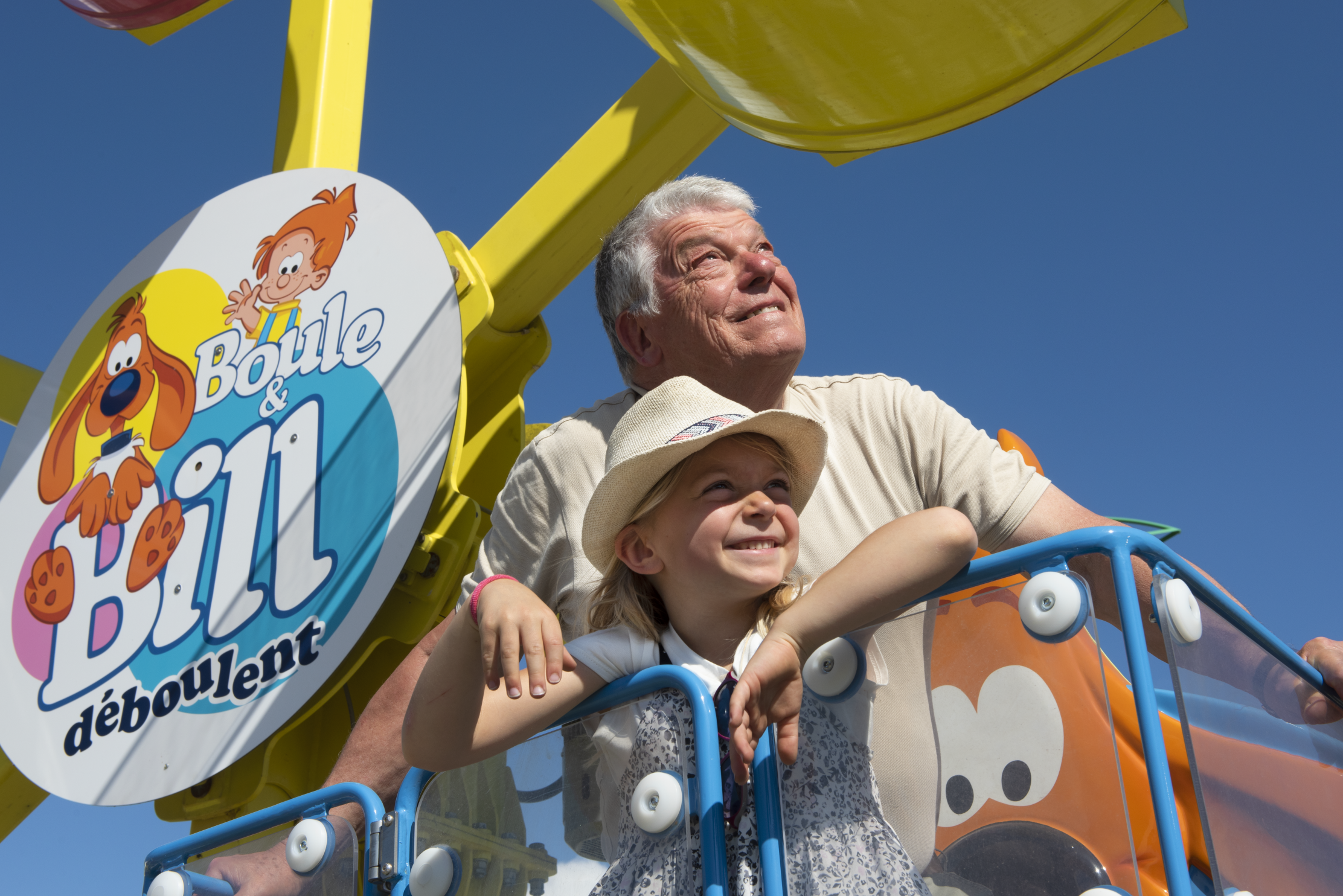 Un homme souriant et une petite fille souriante, portant un chapeau de paille, regardent vers le haut alors qu'ils sont assis dans ce qui semble être un manège d'attraction. Un panneau décoratif avec le texte "Boule & Bill déboulent" et des personnages de dessins animés est visible à gauche.