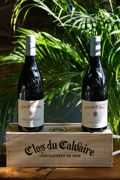 Deux bouteilles de vin sombres avec des étiquettes blanches et des capsules grises, posées sur une caisse en bois clair sur laquelle est inscrit "Clos du Calvaire Châteauneuf du Pape". Un feuillage vert est visible en arrière-plan.