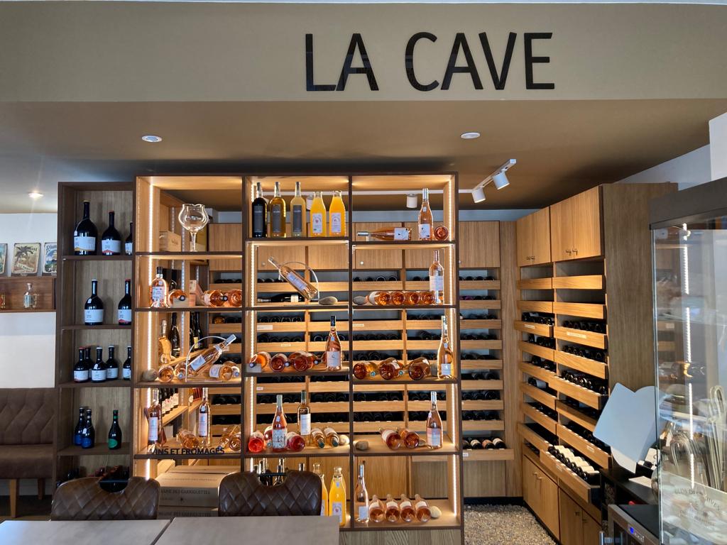 Une vue de l'intérieur d'un magasin de vins et fromages, avec des étagères en bois remplies de bouteilles de vin et de divers fromages et charcuteries dans des vitrines réfrigérées. Le nom "LA CAVE" est visible en lettres noires sur le mur beige au-dessus des étagères.