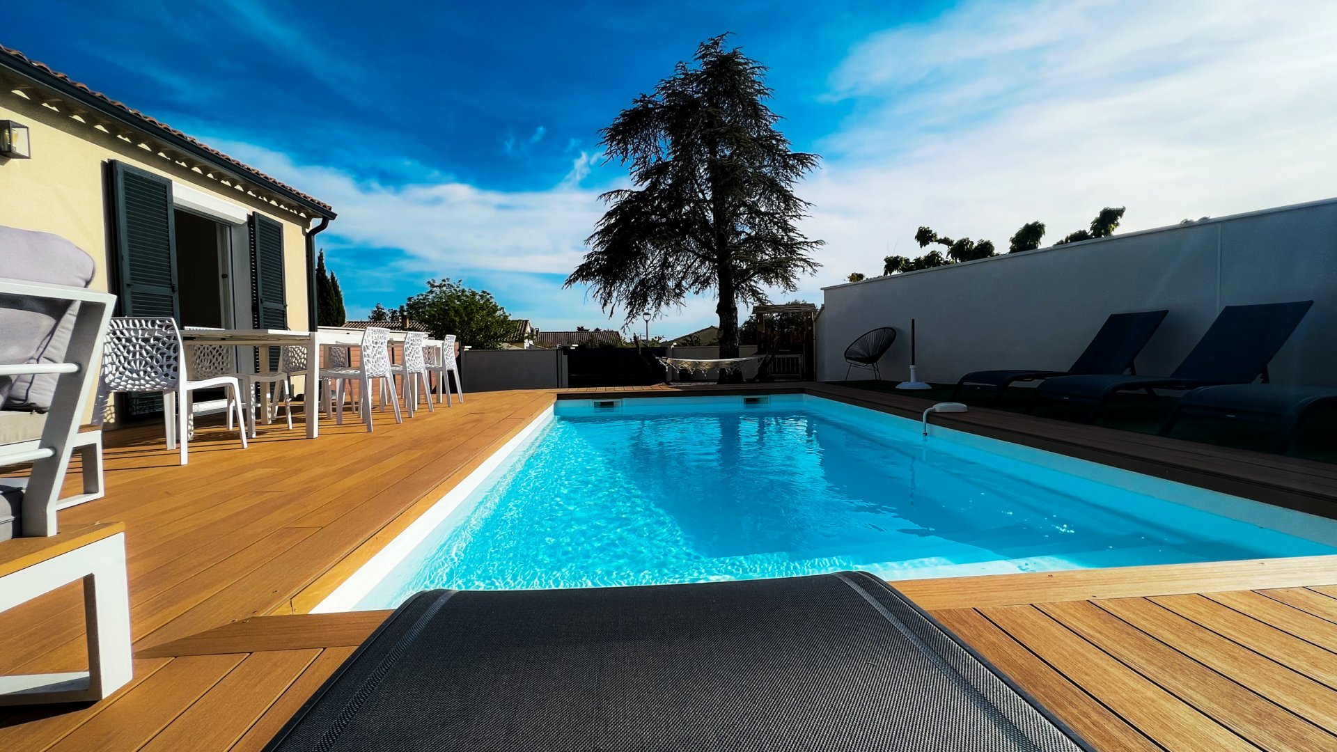 Une terrasse extérieure en bois clair avec une piscine rectangulaire aux eaux bleues. Plusieurs transats sont disposés sur la terrasse, et un ensemble de table et chaises blanches est visible sous une tonnelle. En arrière-plan, on aperçoit une clôture et des arbres, sous un ciel partiellement nuageux.