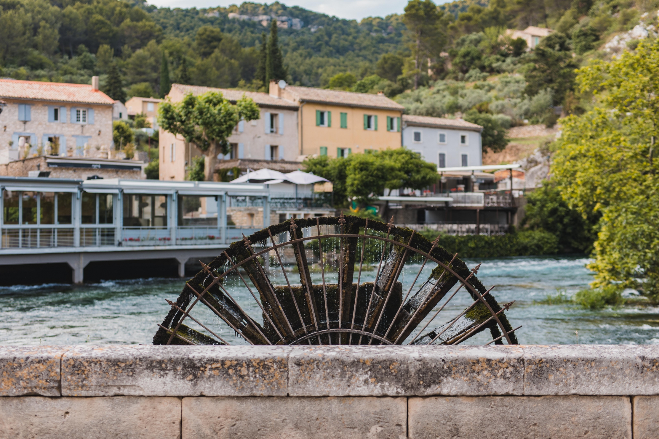 Une roue à aubes en bois émergeant de l'eau devant des bâtiments en pierre et une colline verdoyante à Fontaine-de-Vaucluse, France.