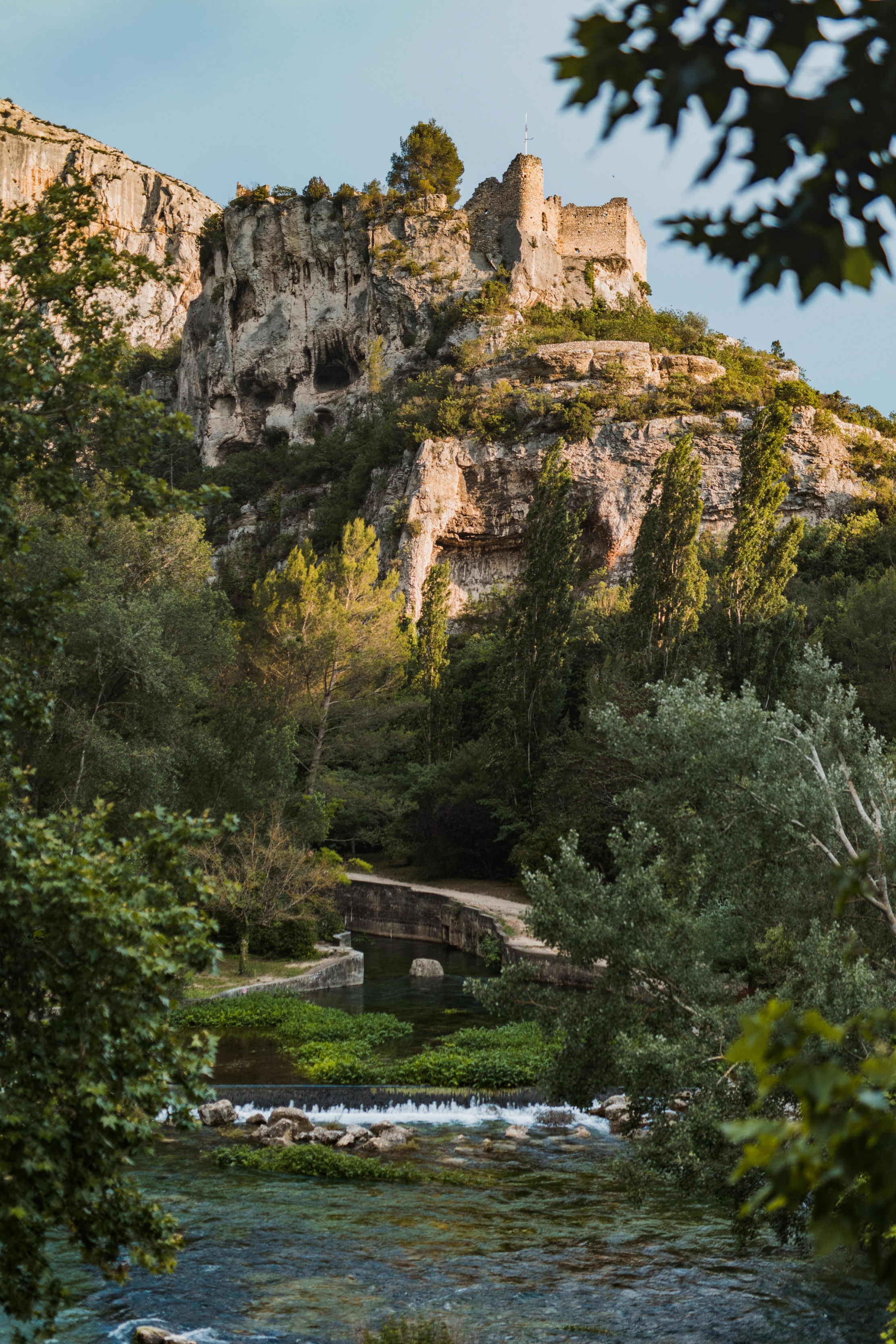 Un ancien château perché sur une falaise rocheuse surplombe une rivière et un pont en pierre, entouré d'arbres luxuriants et de buissons verts à Fontaine-de-Vaucluse.