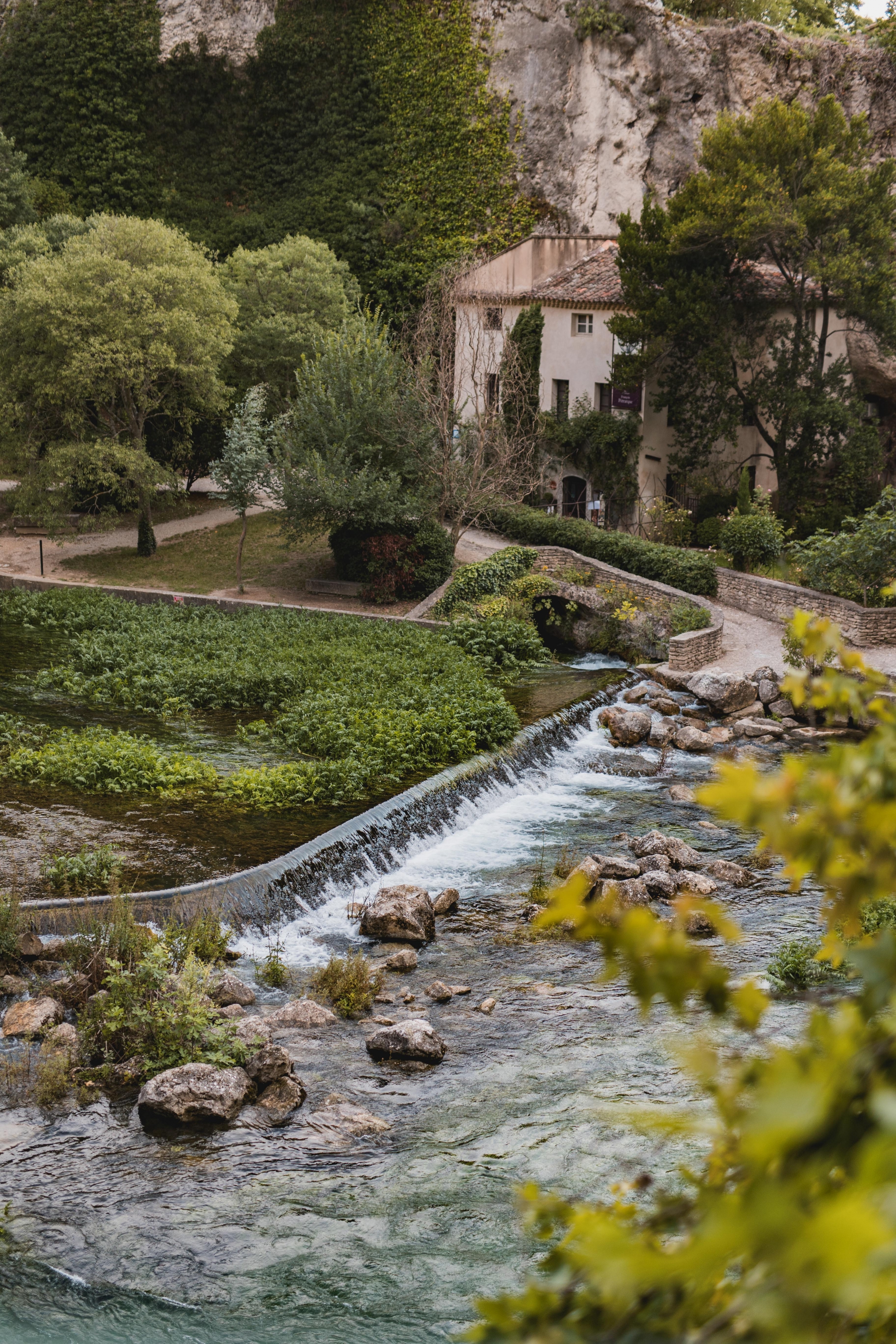 Une vue pittoresque d'une rivière en cascade traversant un village provençal, avec un pont en pierre et une maison ancienne nichée au pied d'une colline verdoyante à Fontaine-de-Vaucluse.