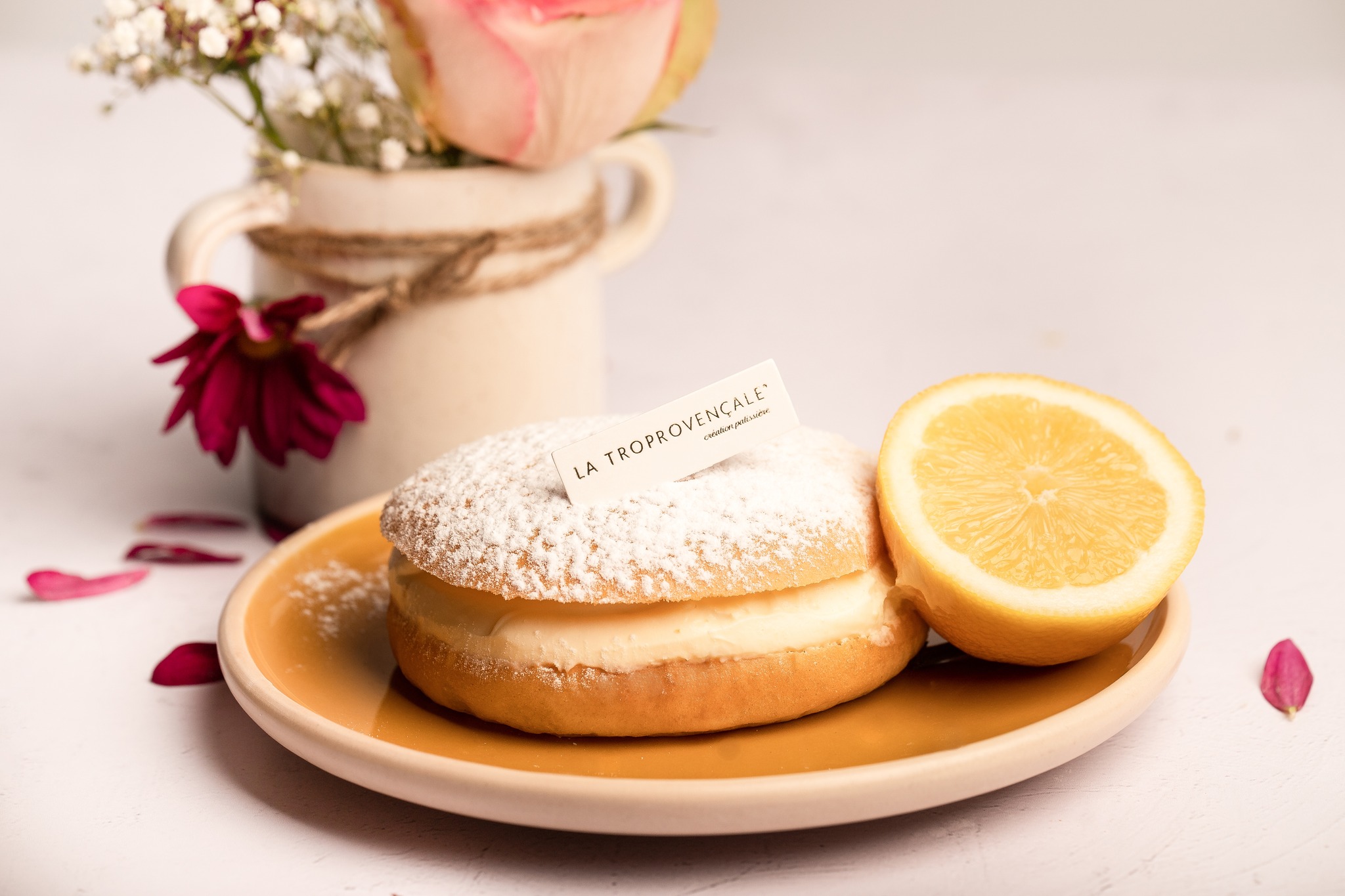 Une pâtisserie ronde et saupoudrée de sucre glace, garnie de crème, est présentée sur une assiette jaune à côté d'un demi-citron. En arrière-plan flou, un vase contient des fleurs blanches et roses avec des pétales rouges éparpillés.