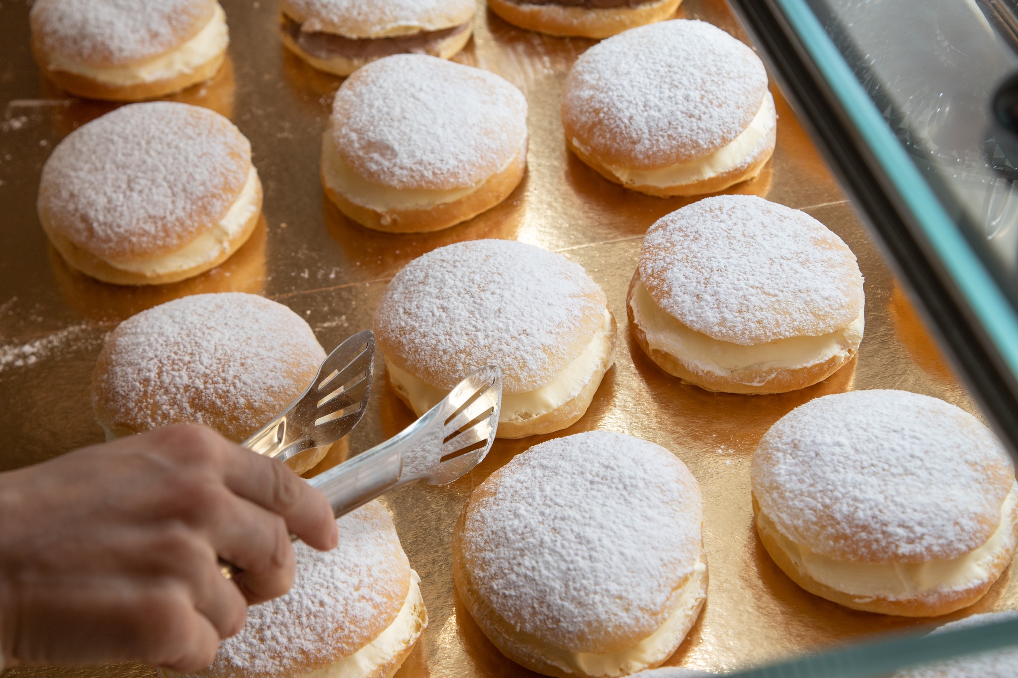 Une main utilise des pinces en métal pour saisir une pâtisserie ronde et saupoudrée de sucre glace, remplie de crème, parmi une douzaine d'autres pâtisseries similaires disposées sur un plateau doré.