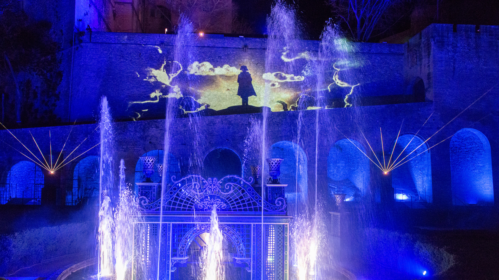 Vue de nuit d'un spectacle lumineux et aquatique projeté sur la façade d'un château. La projection principale au centre montre la silhouette sombre d'une personne debout sur une colline, avec des nuages stylisés en arrière-plan, le tout illuminé en bleu et blanc. Au premier plan, de multiples jets d'eau s'élèvent de fontaines décoratives, éclairées par des lumières bleues vives. Les arches du château et les éléments architecturaux sont également baignés d'une lumière bleue, créant une atmosphère mystique et dramatique.