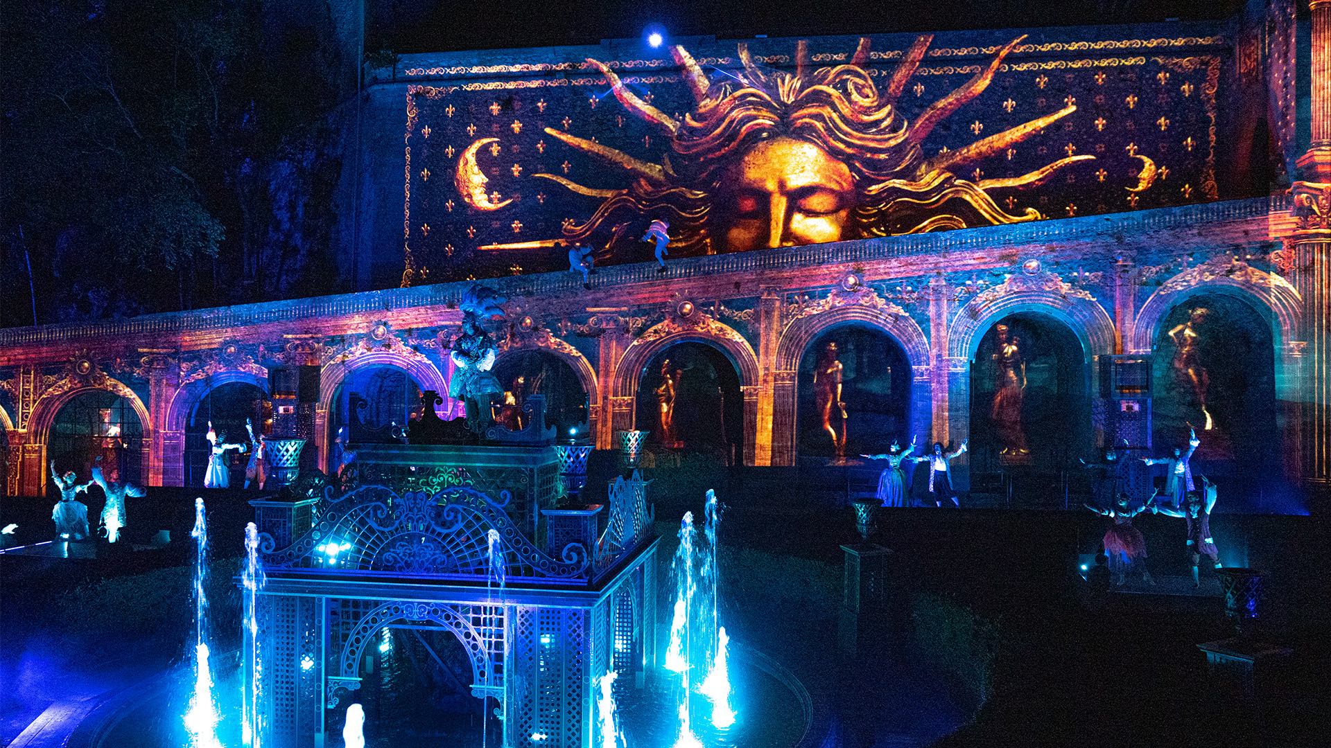 Vue de nuit du château de La Barben, transformé par un spectacle de mapping vidéo appelé 'Les Mystères de la Barben' ou 'La Nuit des Mystères', présentant une projection géante d'un visage doré avec des rayons lumineux au-dessus d'une façade architecturale illuminée. Des arches et des niches abritent des statues et des artistes en costumes blancs, éclairés par des lumières bleues et violettes. Au premier plan, des jeux d'eau illuminés jaillissent de fontaines, ajoutant à l'ambiance féerique. L'ensemble est un spectacle immersif de lumière et de son.