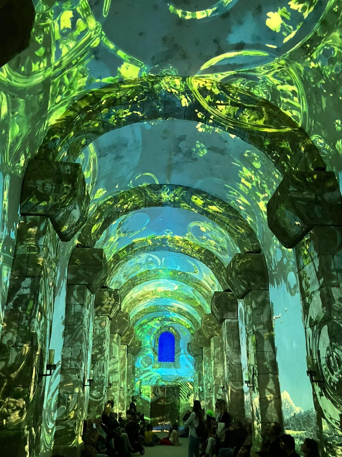 Vue intérieure d'une grande salle voûtée, probablement une église ou une crypte, transformée par une projection immersive de lumières et de motifs végétaux verdoyants. Les murs et les piliers en pierre sont recouverts de motifs abstraits de feuilles et de tourbillons verts et bleus, créant l'impression d'une forêt enchantée. Au fond de la pièce, une fenêtre voûtée est intensément illuminée en bleu vif. Des personnes sont assises sur des bancs ou le sol, observant le spectacle lumineux.