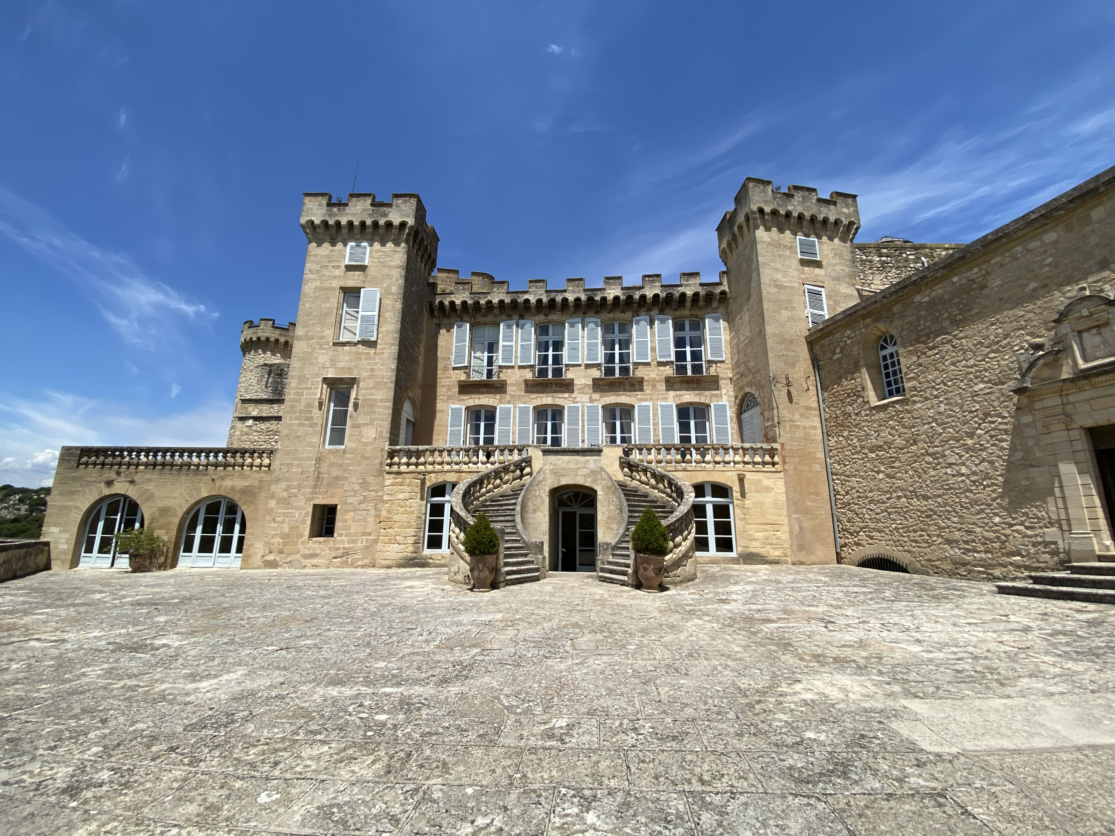 Un majestueux château en pierre de couleur claire avec des tours crénelées et de nombreuses fenêtres aux volets bleus. Un grand escalier à double volée mène à l'entrée principale, et une vaste cour pavée s'étend devant lui sous un ciel bleu clair.