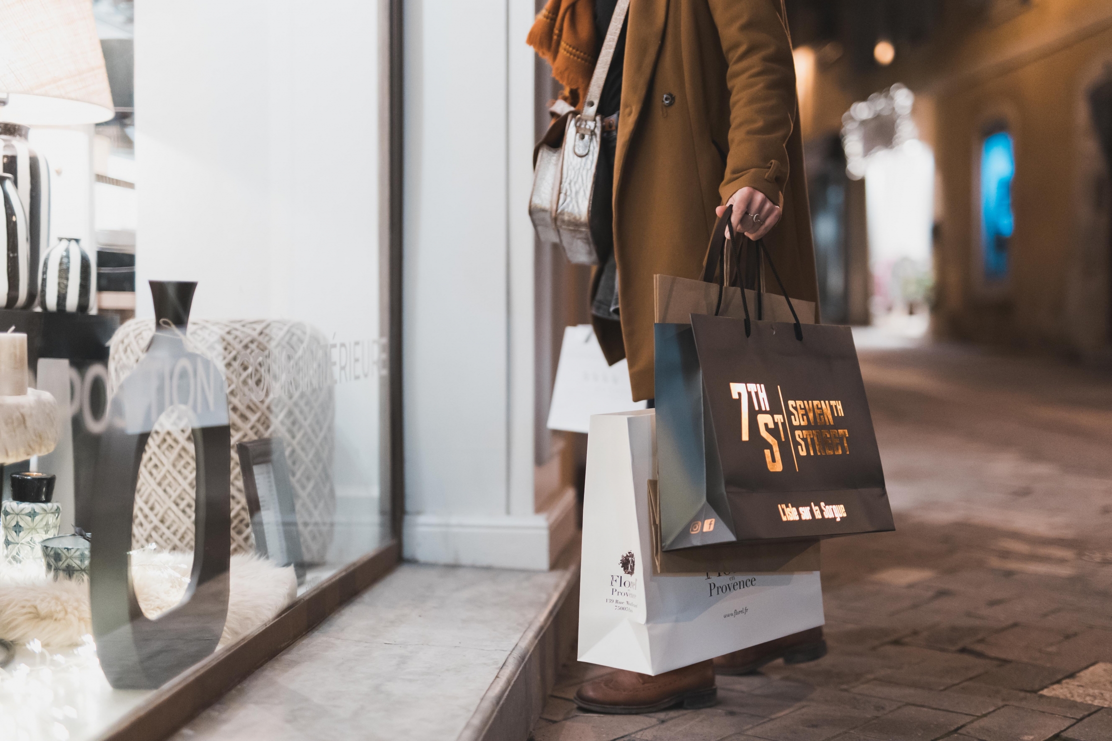 Une personne vêtue d'un long manteau marron, d'un pantalon gris et de chaussures marron, tenant plusieurs sacs de courses, debout devant la vitrine d'un magasin.